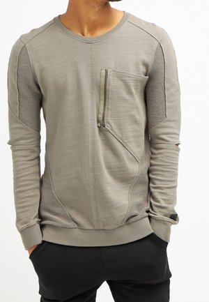Mann trägt hellbraunes langärmeliges Hemd mit Front-Reißverschlusstasche und texturierten Einsätzen, Hände in den Taschen schwarzer Hose, Kopf seitlich gedreht.