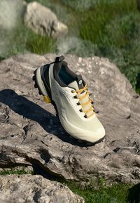 Zapatilla de trail running en color blanco roto con acentos oscuros, cordones amarillos, suela de goma texturizada y marca SCARPA en el lateral.