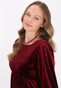 Rochie din catifea roșie, cu mâneci bufante lungi, talie strânsă și un decolteu decorativ legat cu un fir. Textură netedă, cu o culoare bogată și profundă.