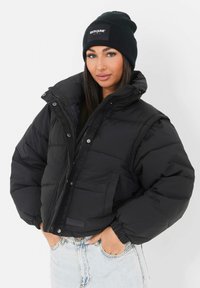 Czarna kurtka puchowa z wysokim kołnierzem, zamkiem błyskawicznym i zatrzaskami, z naszywką z logo i dwoma bocznymi kieszeniami. Ciepły, pikowany fason.