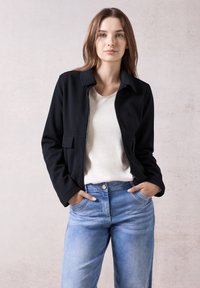 Veste noire à fermeture éclair avec un col, deux poches avant, portée sur un haut de couleur claire, associée à un jean bleu clair. Design minimaliste.