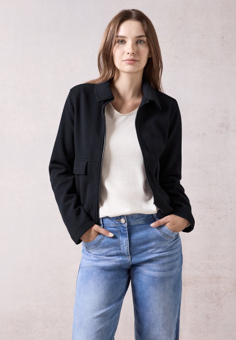 Veste noire à fermeture éclair avec un col, deux poches avant, portée sur un haut de couleur claire, associée à un jean bleu clair. Design minimaliste.