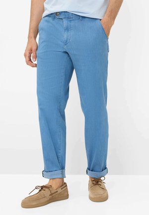 Mand iført lyseblå opkørte bukser med tanfarvede ruskindssko med snørebånd og en hvid skjorte stoppet ind, stående med hænderne i lommerne.