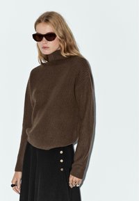 Junge Frau mit blonden Haaren trägt dunkle Sonnenbrillen, einen braunen gerippten Rollkragenpullover und einen schwarzen Rock mit goldenen Knöpfen.