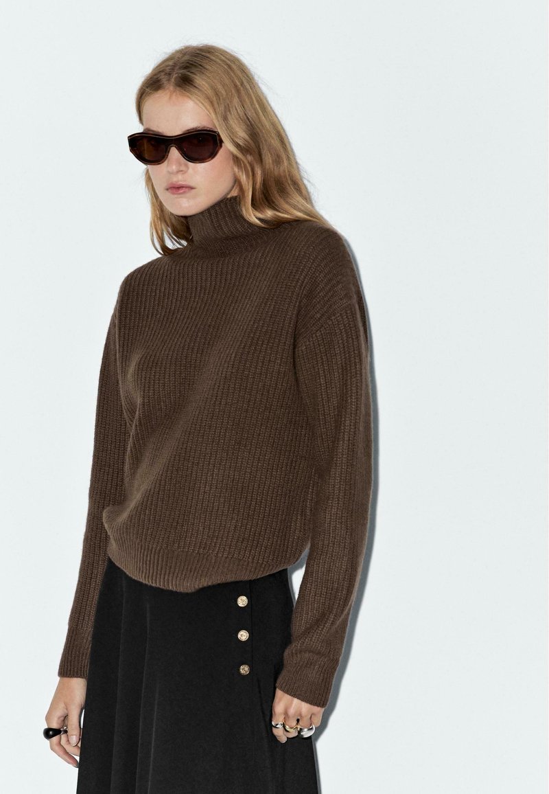 Junge Frau mit blonden Haaren trägt dunkle Sonnenbrillen, einen braunen gerippten Rollkragenpullover und einen schwarzen Rock mit goldenen Knöpfen.