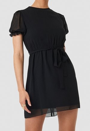 Kvinde iført kort sort kjole med puffede transparente ærmer og taljebånd, stående mod en ensartet lys baggrund.