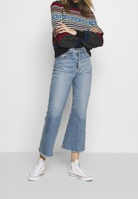 Kvinna står med armarna i kors, iförd en färgglad mönstrad tröja, ljusblå korta jeans och vita högklackade sneakers.