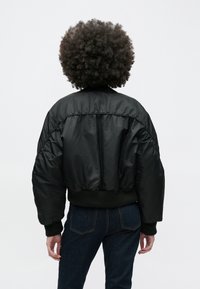 Svart boblejakke med ribbestrikket krage og mansjetter. Har et cropped design og elastisk nederkant. Brukt med mørke jeans, sett bakfra.