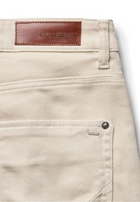 Hlače krem boje s glatkom teksturom, s smeđom kožnom zakrpom na kojoj piše "GABBA", metalnim gumbom i vidljivim detaljima šavova.