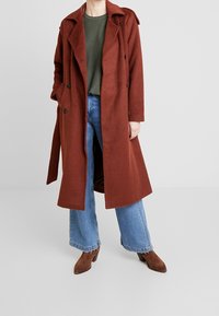 Cappotto di lana color ruggine con un design lungo e sartoriale, doppio petto e grandi tasche, indossato sopra un top verde e jeans a gamba larga blu.