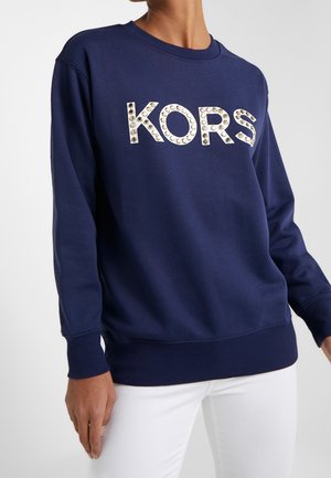 Person iført en marineblå sweatshirt med lange ærmer og ordet "KORS" i store nitter tværs over brystet, kombineret med hvide bukser.