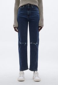 Sötétkék, magas derekú farmernadrág laza szabással, szakadt térdrészletekkel és egyenes szárú kialakítással, fehér sportcipővel párosítva.