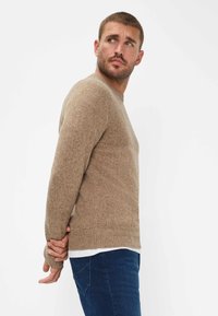 Brązowy dziewiarski sweter z okrągłym dekoltem, teksturowana tkanina i ściągacze na rękawach, zestawiony z ciemnoniebieskimi dżinsami. Model pokazany w profilu bocznym.