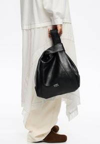 Črna usnjena torba s sproščenim oblikovanjem, ki ima ovito ročico in embosirano logotip. Nosi se z oblačili v svetlih barvah, ki so v plasteh.