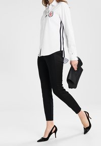 Vrouw in een witte blouse met logo, zwarte slim-fit broek, zwarte slingback hakken, een zwarte clutch tas vasthoudend, aan het lopen.