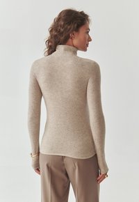 Bež turtleneck-sviiter pehmest kudumisest, millel on pikad varrukad, mugav lõige ja pöidlaharud mansettide juures. Sile tekstuur, minimaalne disain.