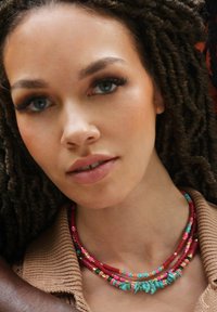 Trois colliers superposés : perles rouges, turquoises et multicolores avec des accents ; portés sur un pull en tricot beige avec un col côtelé.