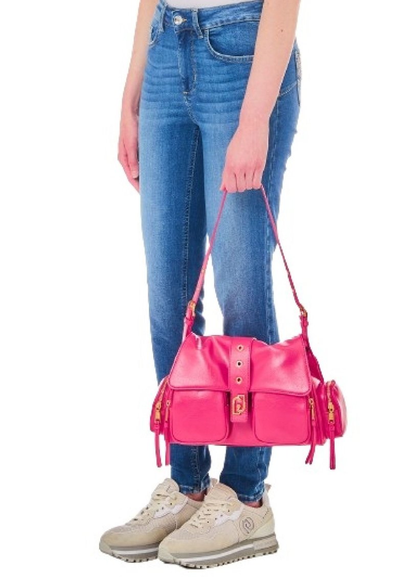 Borsa in pelle rosa con più tasche, dettagli in metallo dorato, tenuta da una persona che indossa jeans blu e scarpe beige. La borsa ha un design strutturato.