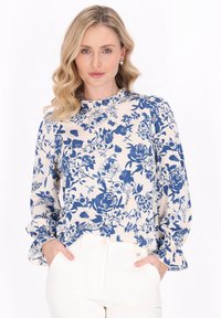 Blusa bianca con motivo floreale blu, scollatura arricciata e maniche lunghe. La modella indossa pantaloni bianchi, mostrando il design e la vestibilità della blusa.