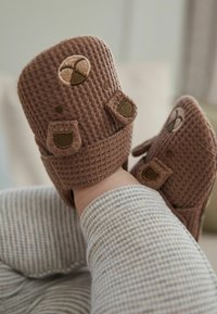Botine maro cu textură de tip vafe, decorate cu aplicații. Prezintă bretele ajustabile și țesătură moale. Combinate cu îmbrăcăminte în dungi gri.