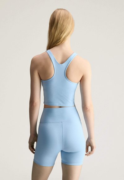 Femme aux longs cheveux blonds portant un crop top bleu clair dos nageur et un short cycliste taille haute assorti, debout devant un fond neutre.