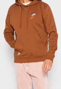 Sudadera marrón de Nike con bolsillo canguro y capucha con cordón. Presenta múltiples logotipos bordados en blanco y naranja.
