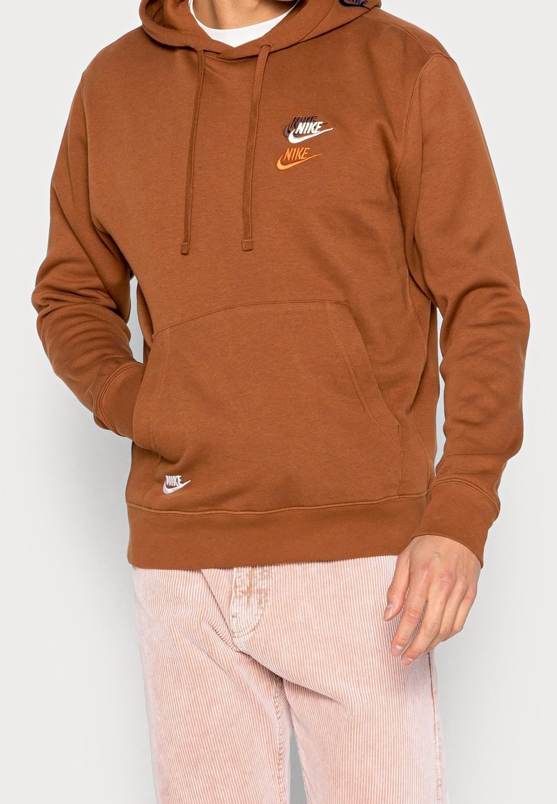 Sudadera marrón de Nike con bolsillo canguro y capucha con cordón. Presenta múltiples logotipos bordados en blanco y naranja.
