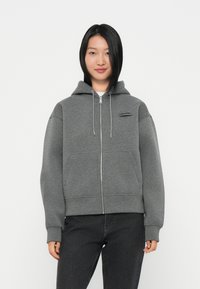 Pilkas užtrauktukas su kapišonu, turintis priekines kišenes ir mažą juodą logotipą ant krūtinės. Audinys atrodo minkštas ir tekstūruotas.