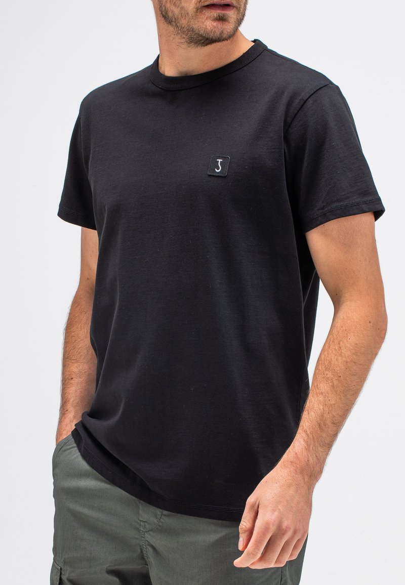 Schwarzes Baumwoll-T-Shirt mit Rundhalsausschnitt, kurzen Ärmeln und einem kleinen quadratischen Logo auf der linken Brust. Glatte Textur, lässige Passform.