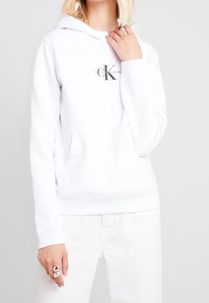 Woman wearing white Calvin Klein Jeans hoodie with front pocket and white pants on light gray background, hand near face.