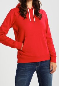 Czerwona bluza z kapturem wykonana z gładkiego materiału, z przednią kieszenią, białymi sznurkami i ściągaczami przy mankietach oraz u dołu. Noszona z niebieskimi dżinsami.