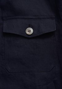 Navy linen fabric with a front chest pocket featuring a silver button and contrasting stitching, providing a textured appearance.