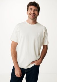 Bílé tričko s krátkými rukávy a kulatým výstřihem, se malou čtvercovou nášivkou na hrudi, v kombinaci s tmavě modrými džíny. Hladká textura.