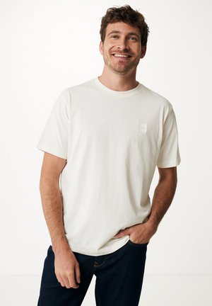 Camiseta blanca de manga corta con cuello redondo y pequeña etiqueta cuadrada en el pecho, combinada con jeans azul oscuro. Textura suave.
