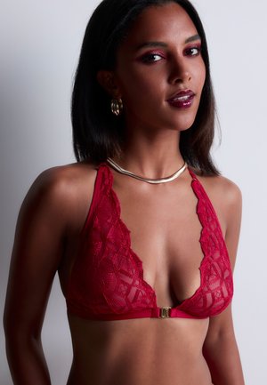 Femme à la peau moyenne foncée portant un bralette en dentelle rouge, un collier en or et des boucles d'oreilles créoles, posant sur un fond neutre.