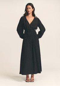 Black polka dot maxi dress with a deep V-neck, long puff sleeves, and an elastic waistband. Lightweight fabric drapes elegantly to the floor.