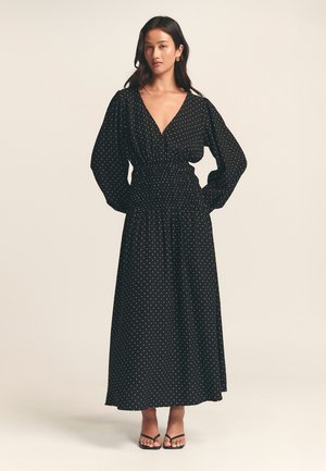 Black polka dot maxi dress with a deep V-neck, long puff sleeves, and an elastic waistband. Lightweight fabric drapes elegantly to the floor.