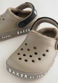 Zoccoli beige con interno morbido foderato in pelliccia, molteplici fori di ventilazione e una cintura nera. Marchio "BAKER" sul lato e sul tallone.