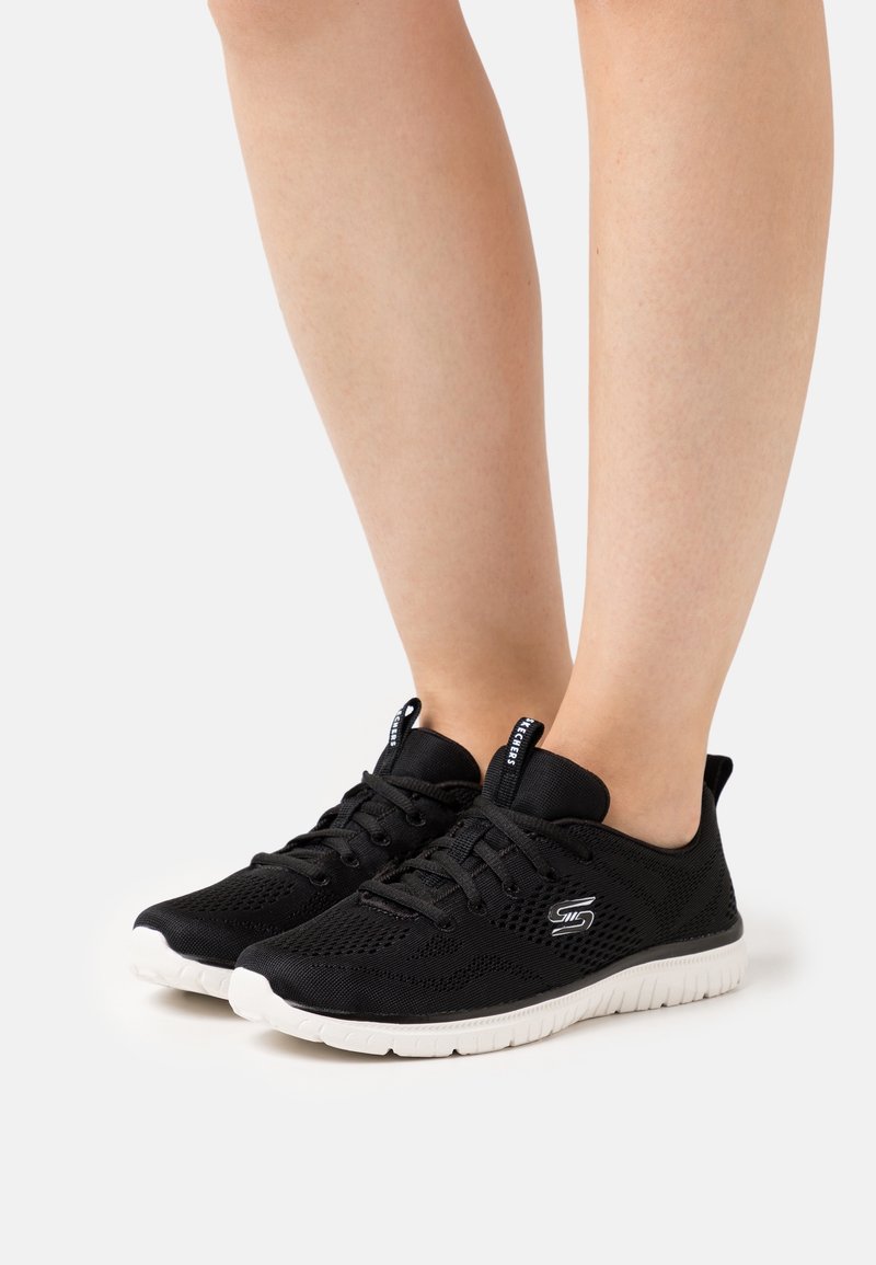 Baskets légères en mesh noir avec semelles blanches portées aux pieds, comprenant un design à lacets et le logo Skechers sur les côtés.