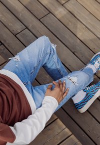 Vaqueros de mezclilla azul claro desgastados con roturas, combinados con una camiseta blanca y marrón. El calzado presenta un diseño de patrones ondulados vibrantes en azul y naranja.