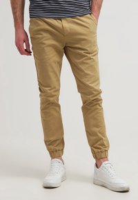 Man som bär beige joggarbyxor med elastiska muddar, vita sneakers och en svart-vit rutig skjorta, stående med en hand i fickan.