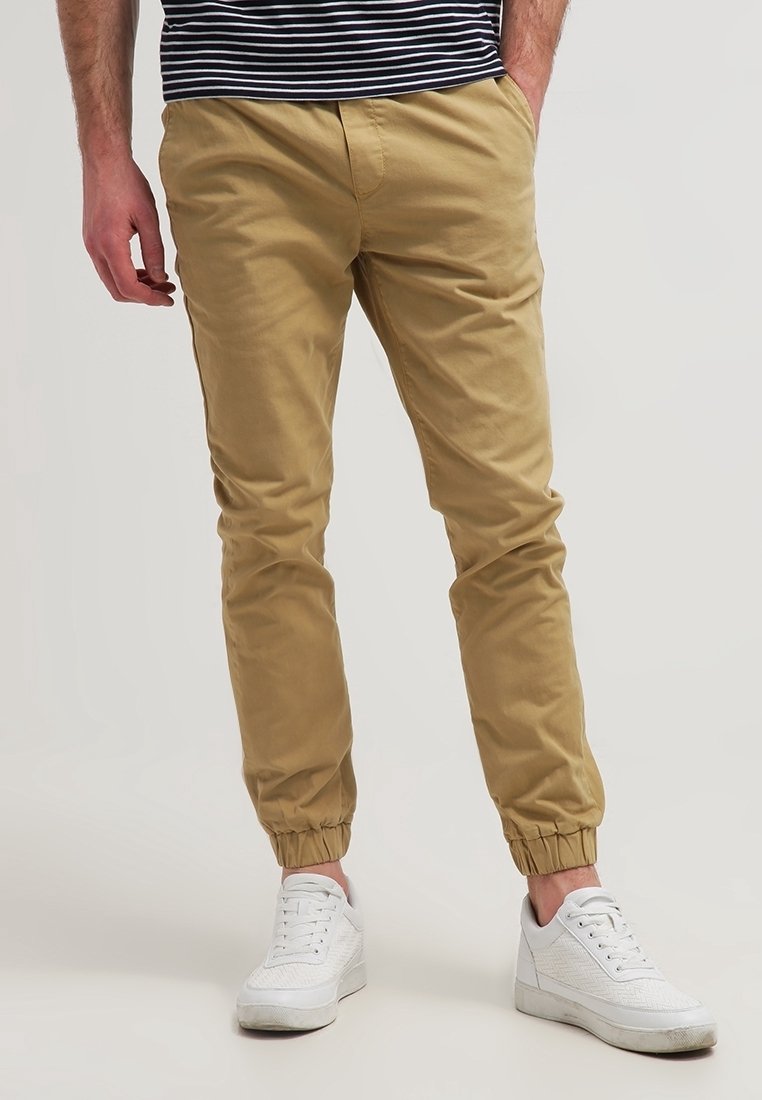 Man som bär beige joggarbyxor med elastiska muddar, vita sneakers och en svart-vit rutig skjorta, stående med en hand i fickan.