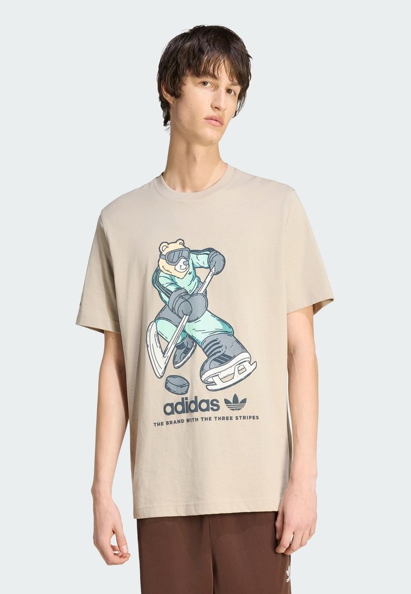 Maglietta beige in cotone con grafica di un orso in tenuta sportiva, che tiene una mazza da hockey. Logo "adidas" e testo stampati sotto il disegno.