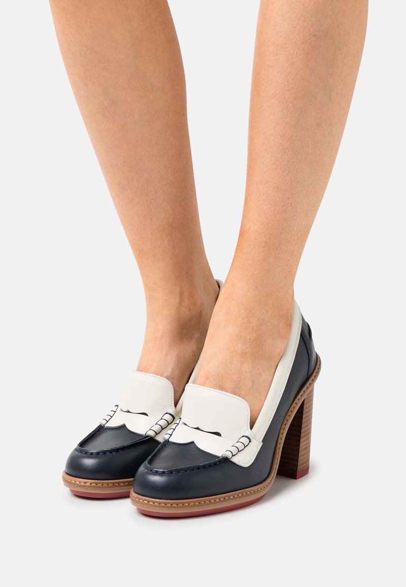 Pieds portant des mocassins à talons hauts en cuir noir et blanc avec des talons carrés en bois et des semelles cousues marron sur un fond blanc.