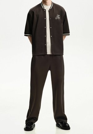 Persona que lleva una chaqueta marrón oscuro de manga corta con botones sobre una camisa blanca, pantalones a juego y zapatos negros sobre un fondo blanco liso.