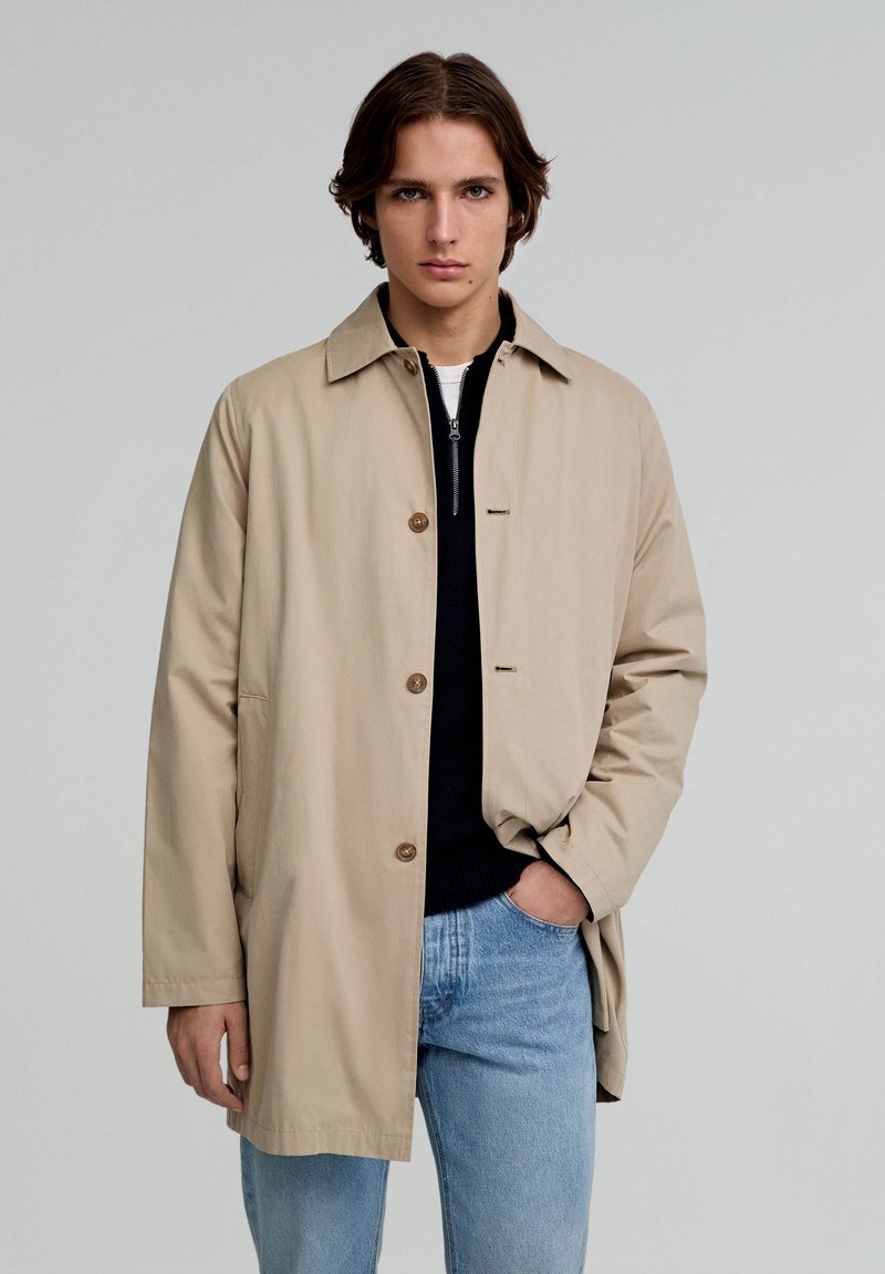 Beige, lichtgewicht jas met een knoopsluiting, een kraag en twee zijzakken. Draag het over een donkere trui en lichte spijkerbroek.