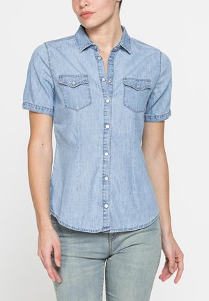 Donna che indossa una camicia di jeans azzurro chiaro a maniche corte con bottoni frontali e tasche sul petto, abbinata a jeans di jeans azzurro chiaro.
