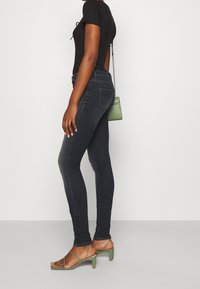Jeans ajustados de un azul oscuro, confeccionados en denim, combinados con una blusa negra de canalé y sandalias verdes de tacón. La figura lleva un pequeño bolso verde.