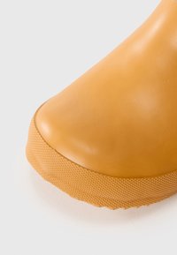 Botte en caoutchouc avec une surface lisse et mate, un bout arrondi, et une semelle en forme de coin texturée de couleur jaune moutarde.
