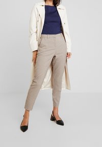 Beige rutiga byxor med en smal passform och hög midja, parat med en marinblå topp och en ljus gräddfärgad kappa, kompletterat med svarta klackar.